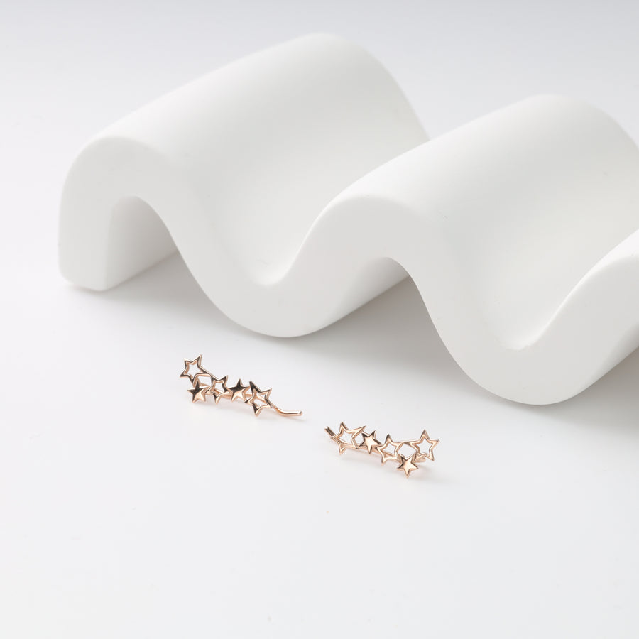 Pair of rose gold star ear climbers with five hollow stars each on a white wavy display, rose gold earrings.