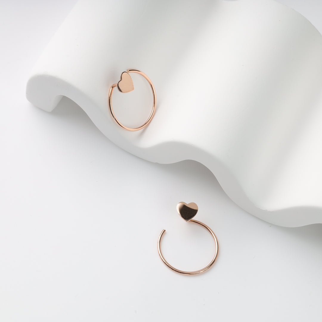 Two rose gold open hoop earrings with heart ends on a white wavy display prop, rose gold hoop earrings, heart hoop earrings.