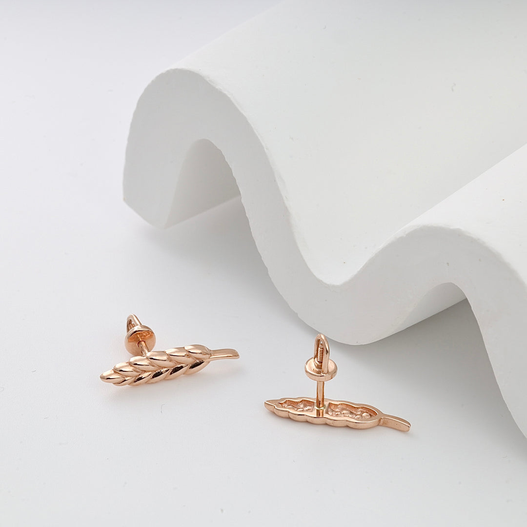 Rose gold stud earrings with a wheat leaf design displayed on a white curved backdrop.
