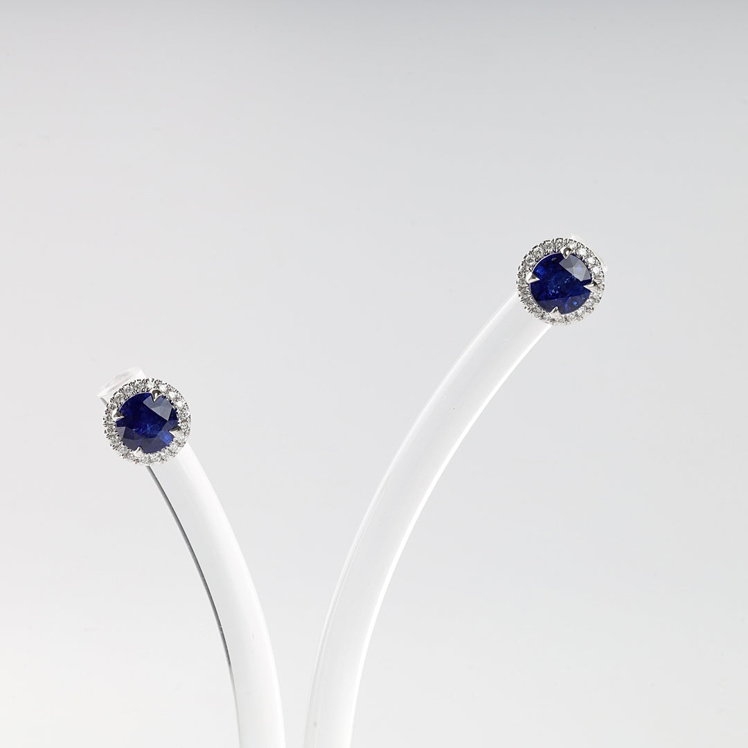 Pair of round sapphire stud earrings with a halo of clear stones, set in silver, displayed on a white stand.