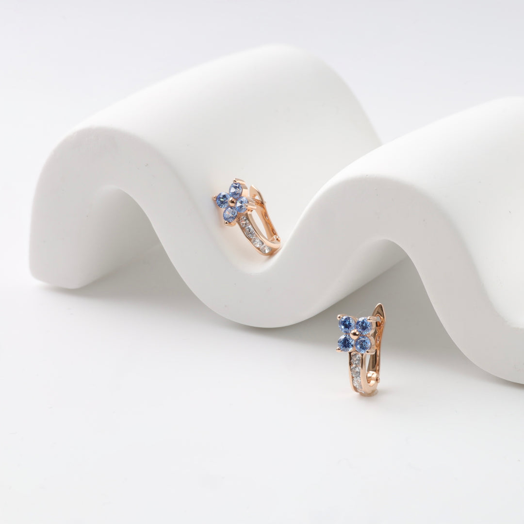 Gold hoop earrings with blue gemstone flowers on a white wavy background.