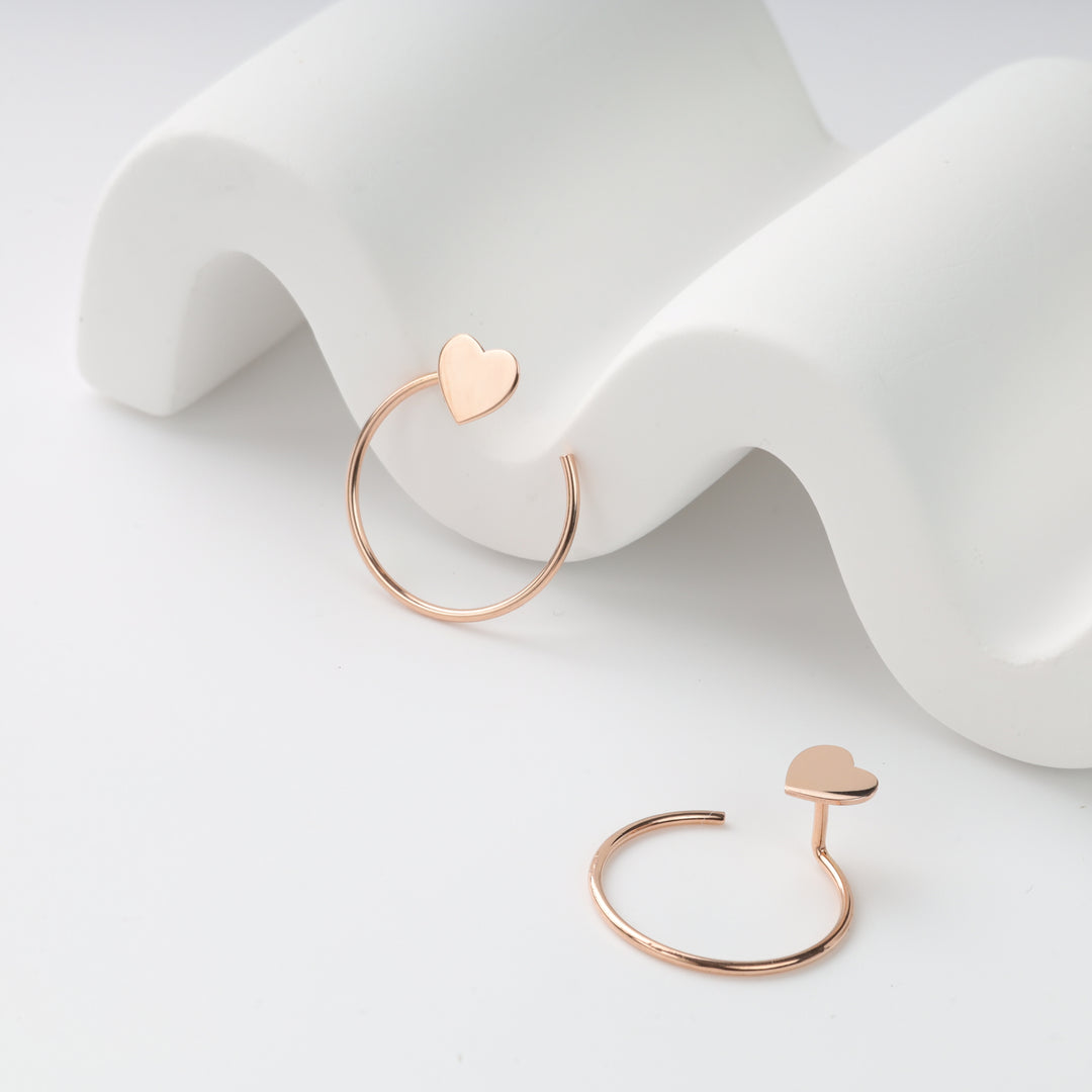 Pair of rose gold open hoop earrings with heart studs displayed on a white wavy prop, rose gold hoop earrings, heart stud hoops.