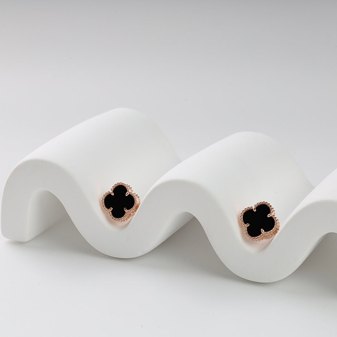 Pair of black enamel four-leaf clover stud earrings with gold trim displayed on a white wavy stand.