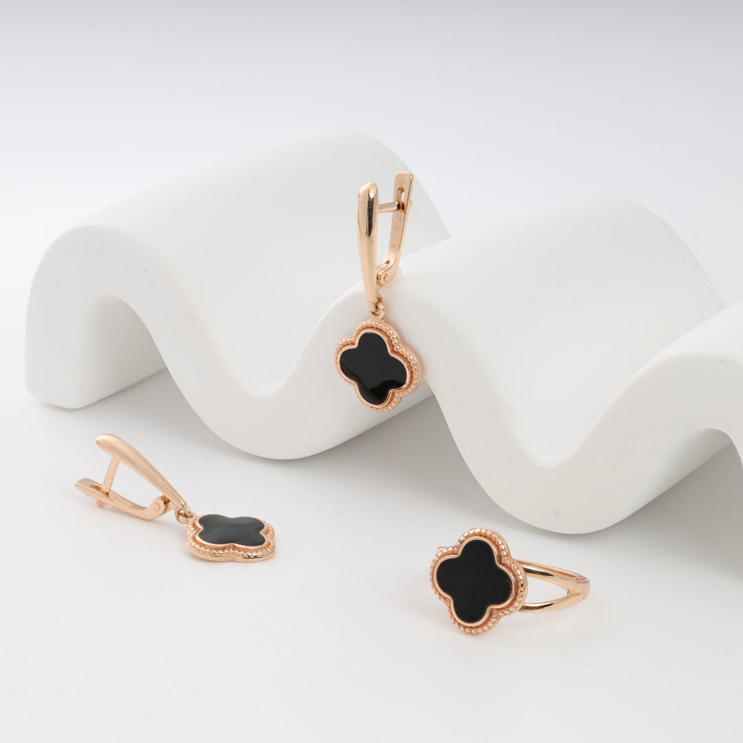 Rose gold earrings and ring set with black clover motifs displayed on a white wavy surface.