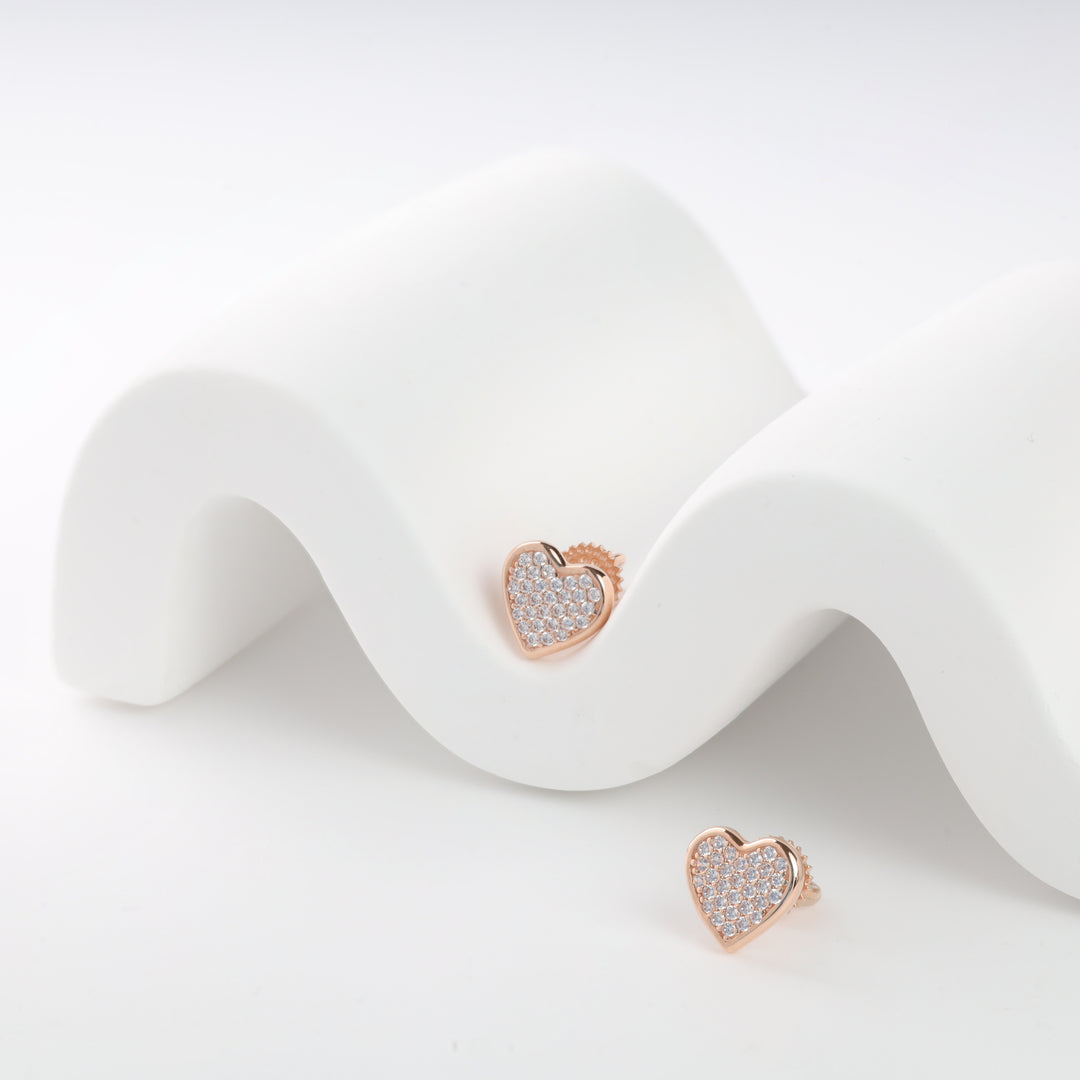 Rose gold heart-shaped stud earrings with crystal embellishments on a white wave-pattern display.