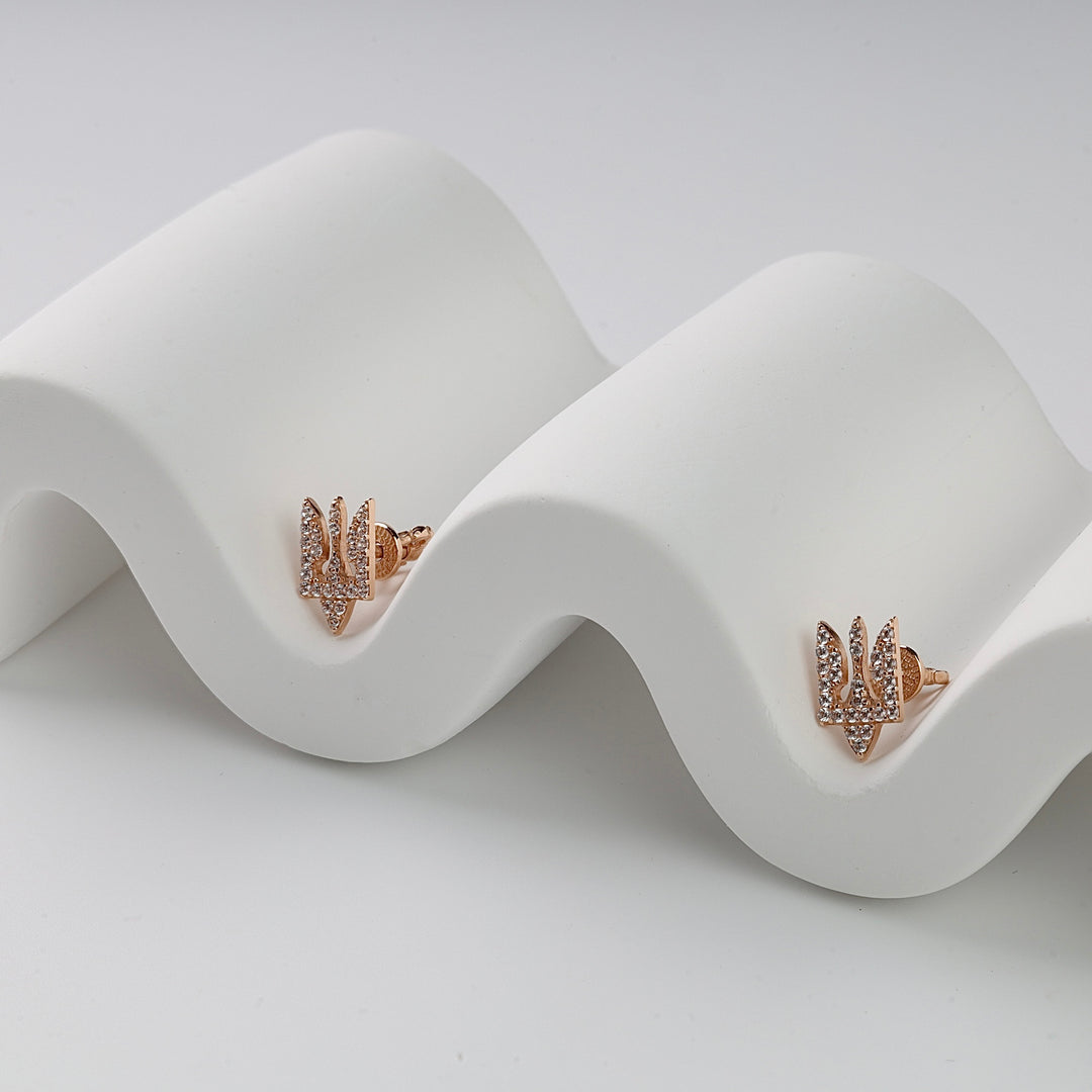 Gold trident stud earrings with clear crystal accents displayed on a white wavy surface, Ukraine jewelry, dainty studs.