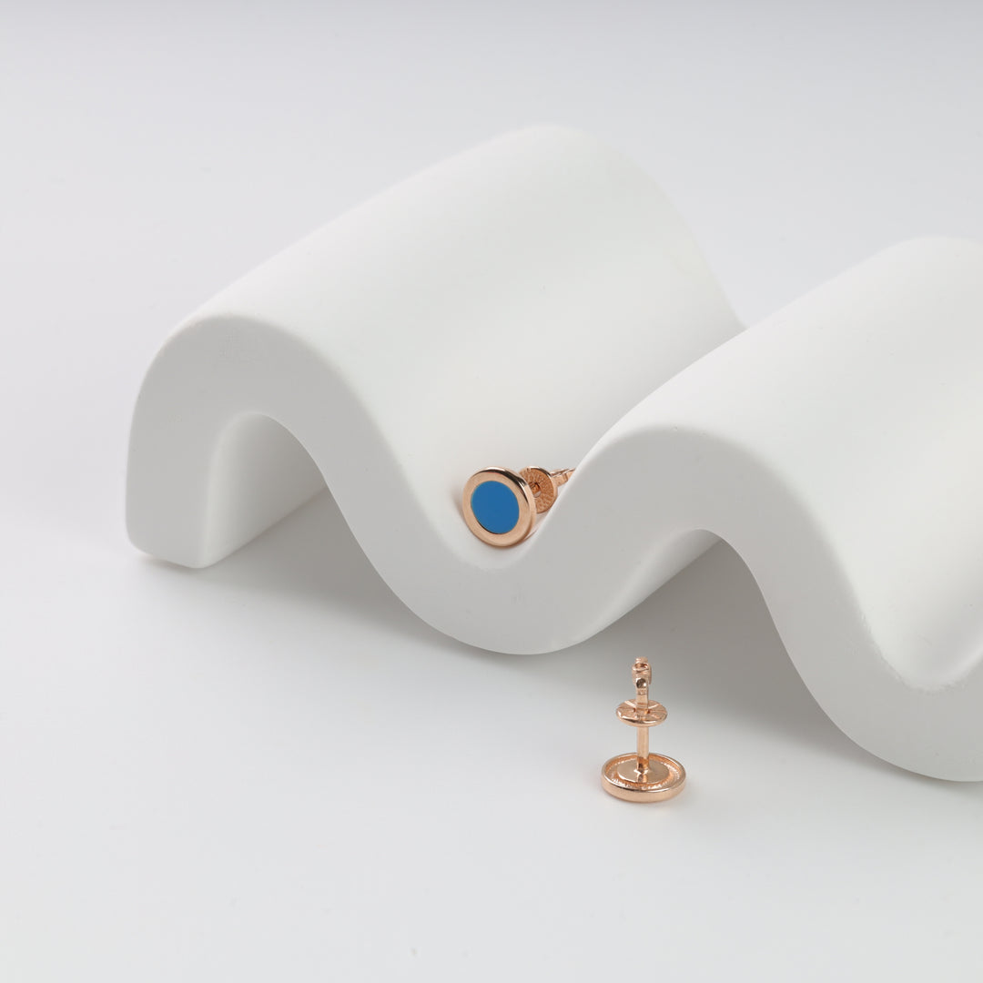 Pair of round gold stud earrings with blue enamel center, displayed on a white wavy stand, modern jewelry.