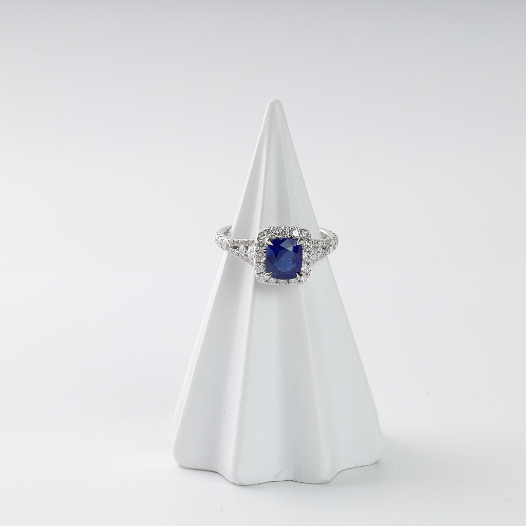 Silver ring with a cushion-cut blue gemstone and clear accent stones displayed on a white cone stand.