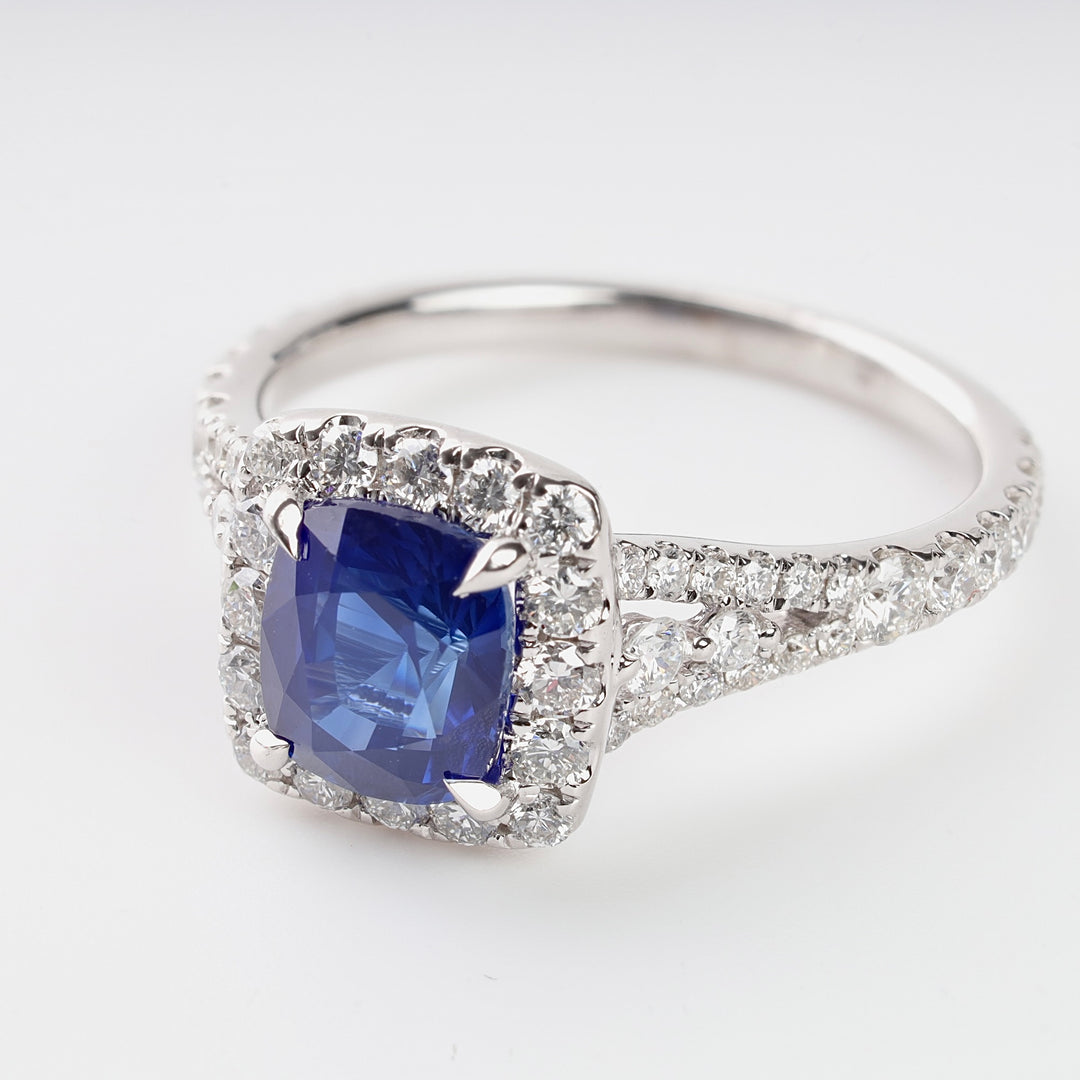 White gold ring with a cushion-cut blue sapphire center stone and a halo of round white diamonds, shown on a plain background.