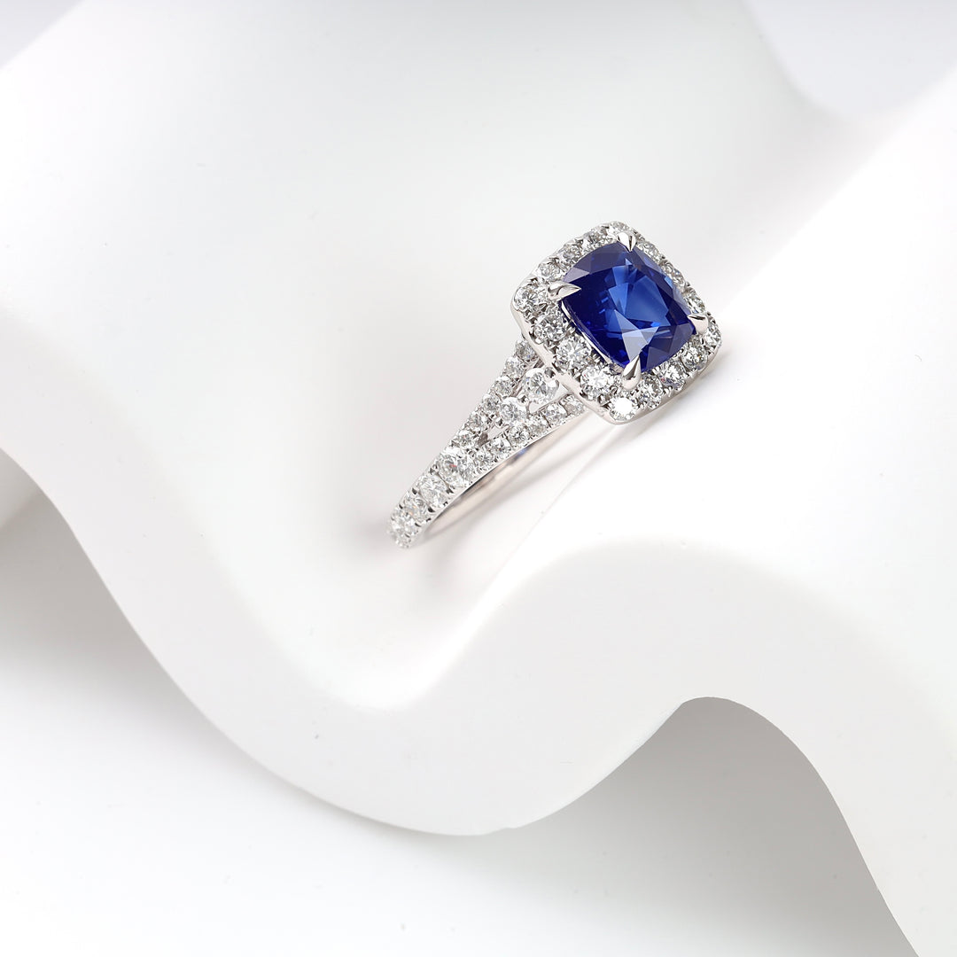 White gold ring with cushion-cut blue sapphire and pavé diamond band displayed on a white wavy surface.