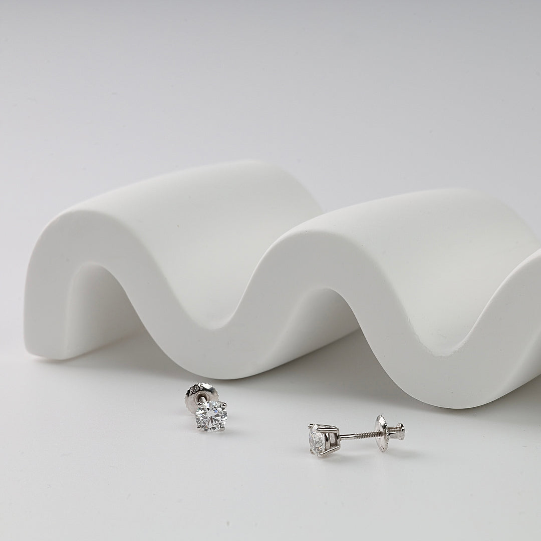 Pair of white gold stud earrings with round clear gemstones shown on a white wave-shaped display.