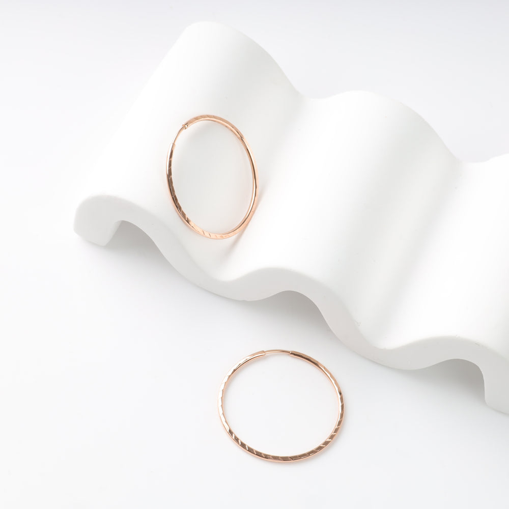 Two rose gold hoop earrings displayed on a white, wavy surface.