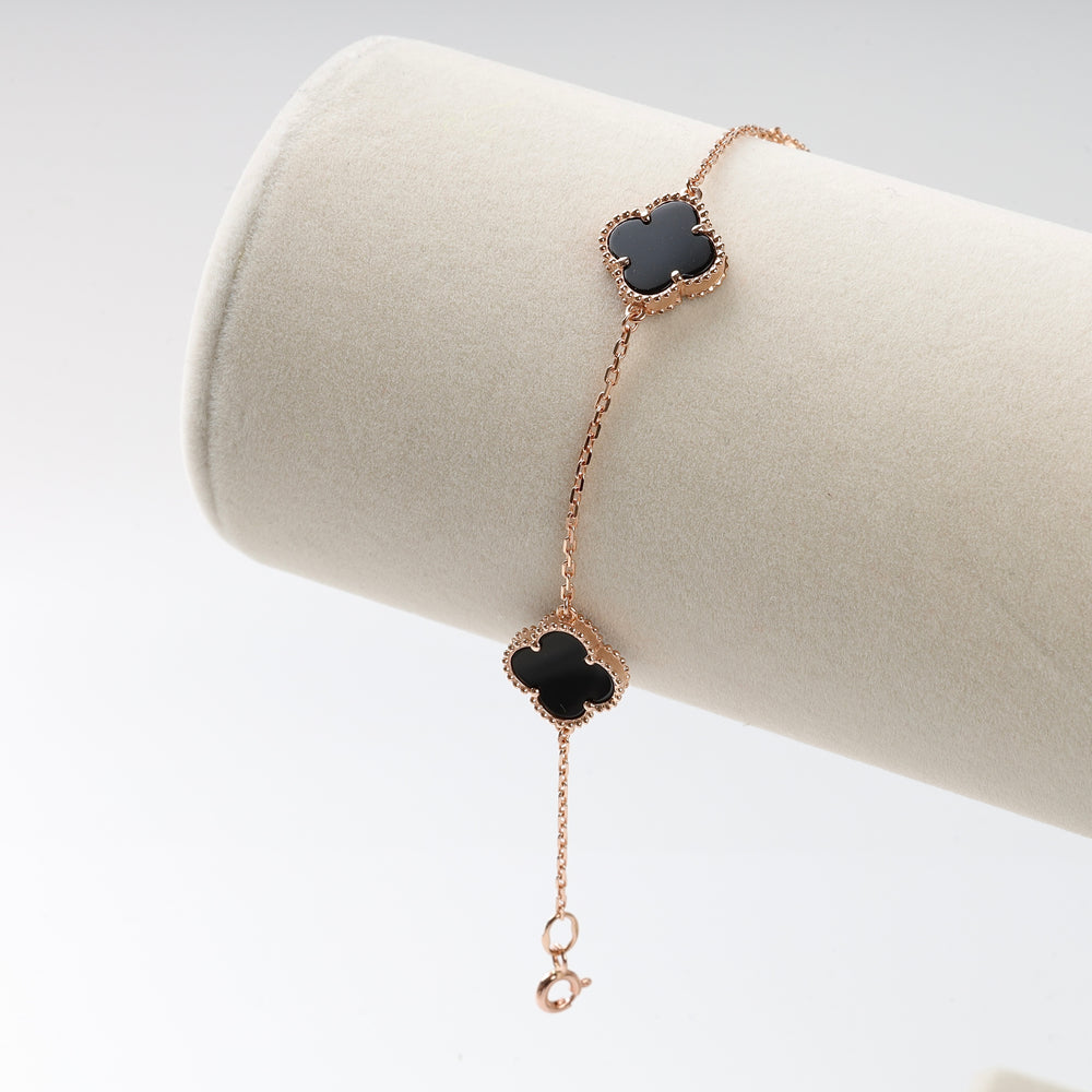 Rose gold chain bracelet with two black clover shaped charms displayed on a cream jewelry roll.