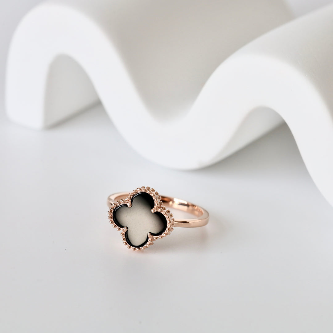 Rose gold ring with a black clover-shaped enamel centerpiece shown on a white background.