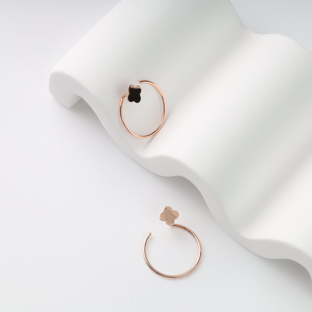 Two rose gold hoop earrings with clover charms (one black, one rose gold) on a white wavy display, rose gold hoops, clover earrings.