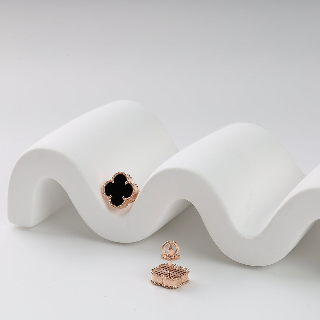 A pair of rose gold clover stud earrings with black enamel displayed on a white wavy prop.