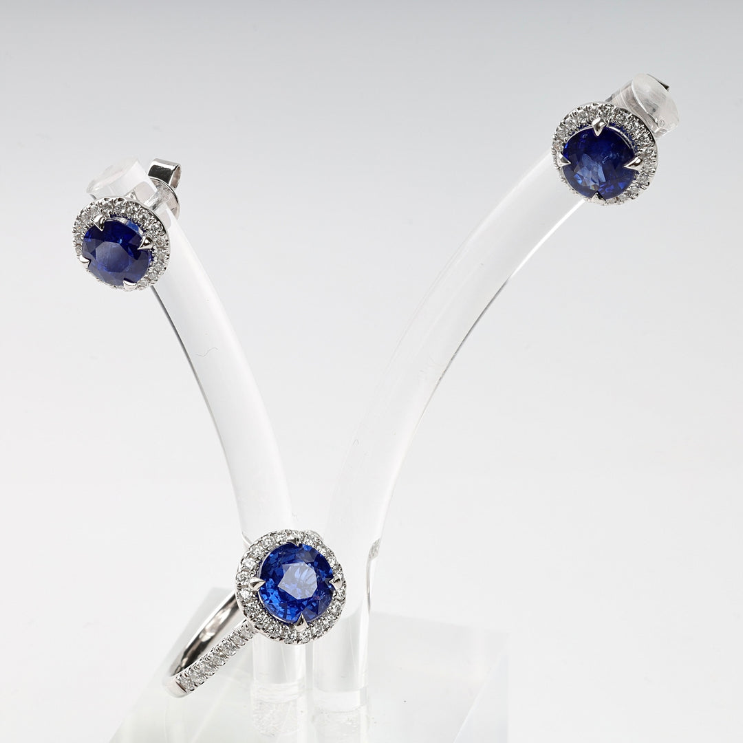 Silver ring and stud earrings set with round blue stones and clear stone halos on a clear display stand. Sapphire jewelry set.