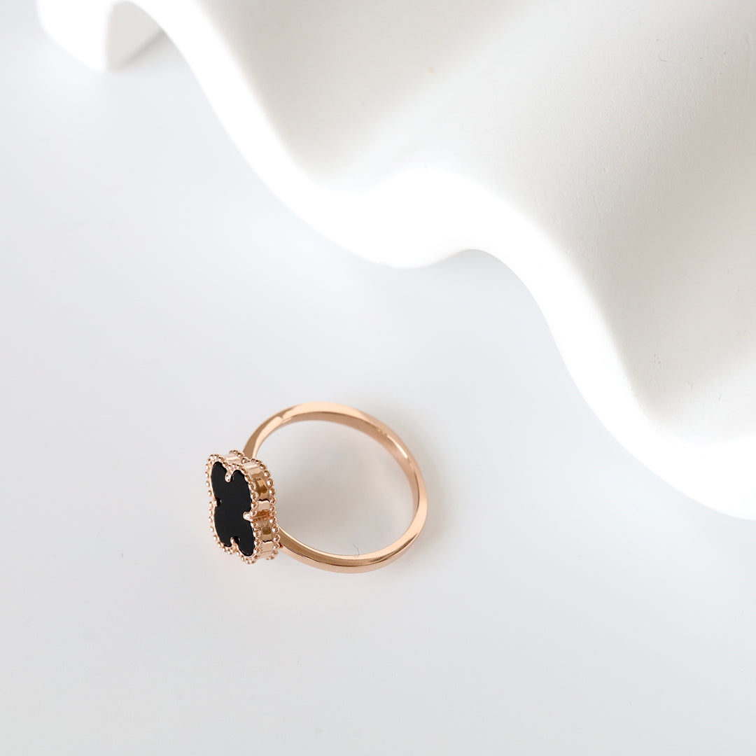 Gold ring with a black clover-shaped stone and textured edge displayed on a white surface. Gold clover ring, black gemstone.