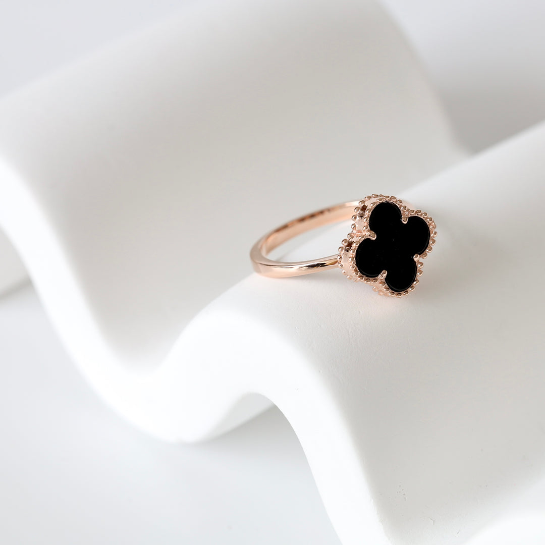 Gold ring with black clover-shaped enamel face displayed on a white curved stand. Gold ring, black enamel, clover motif.