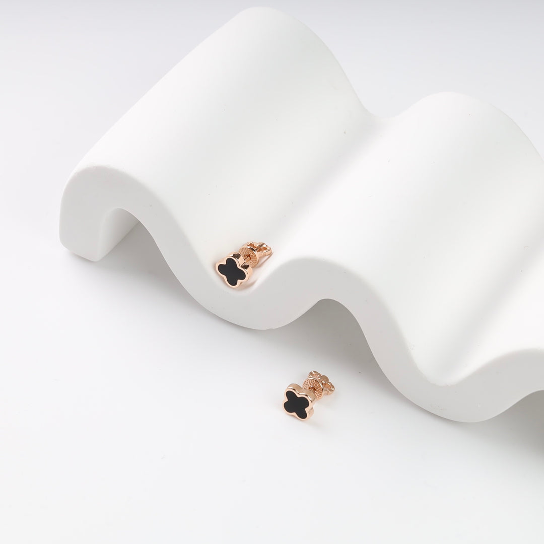 Black clover stud earrings with rose gold trim displayed on a white wave surface.