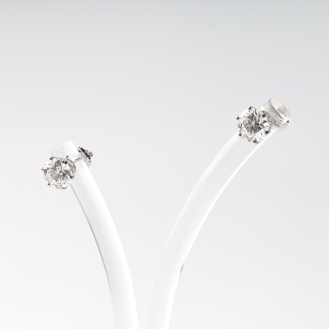 Pair of round white diamond stud earrings in silver settings displayed on a clear acrylic stand.