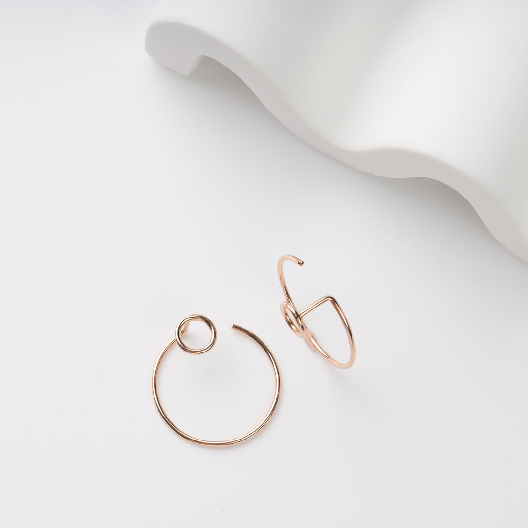 Two rose gold wire earrings on white surface, one open circle hoop and one angular loop; minimalist hoop earrings, geometric earrings.