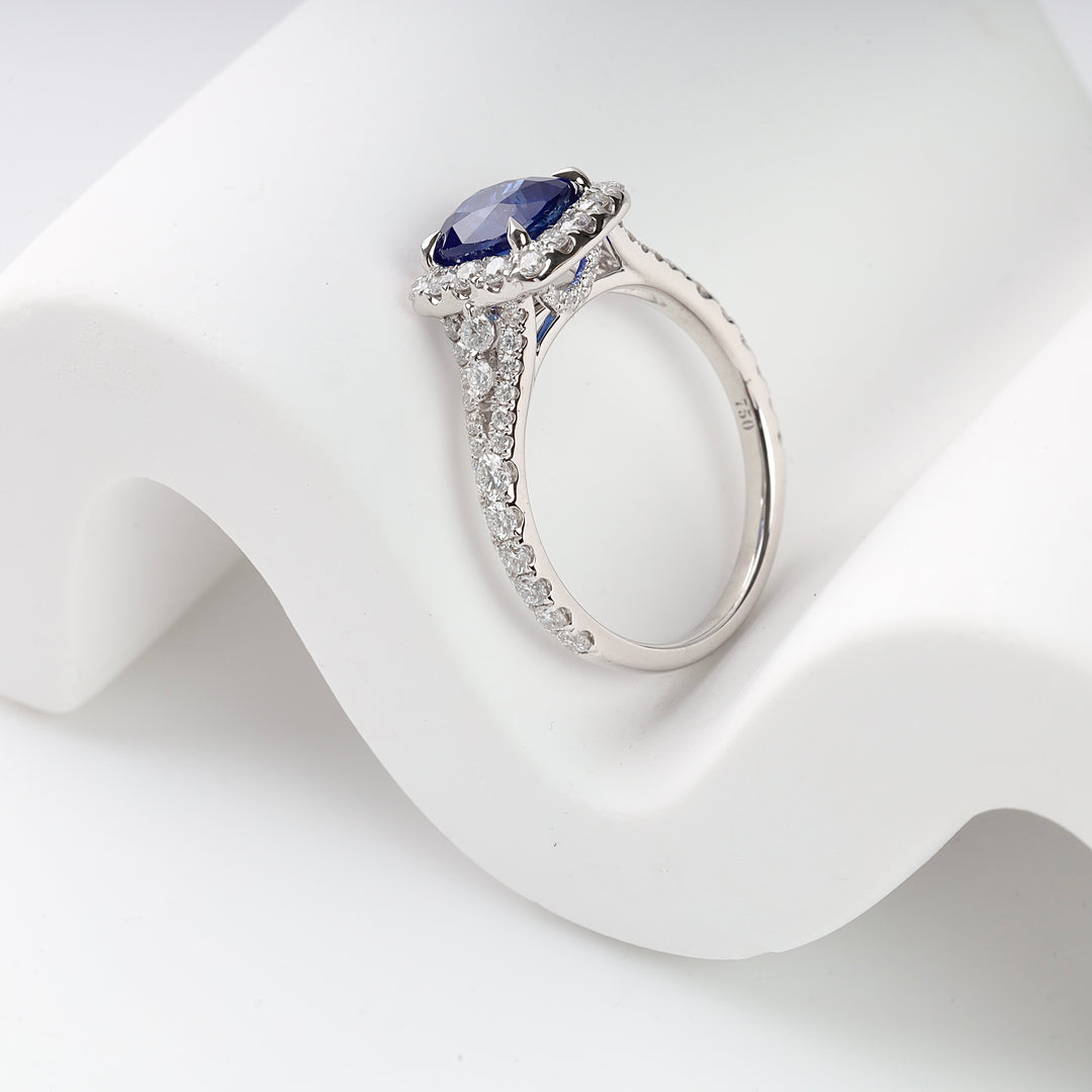 White gold ring with a large oval blue sapphire and small clear diamonds on band, displayed on a white curved surface.