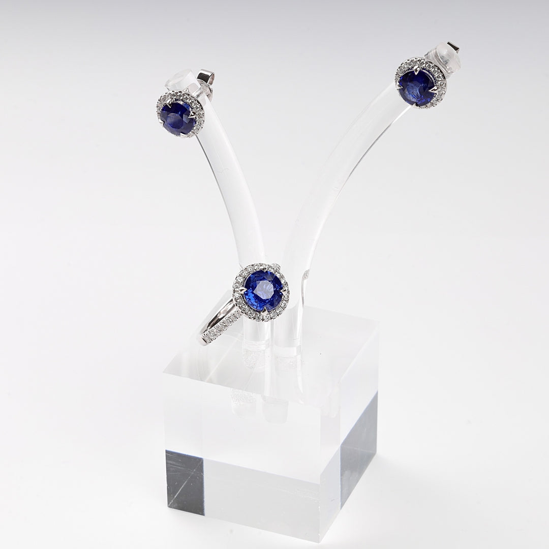 Silver ring and stud earrings set with round blue stones and clear accents displayed on a clear stand. Sapphire jewelry set.