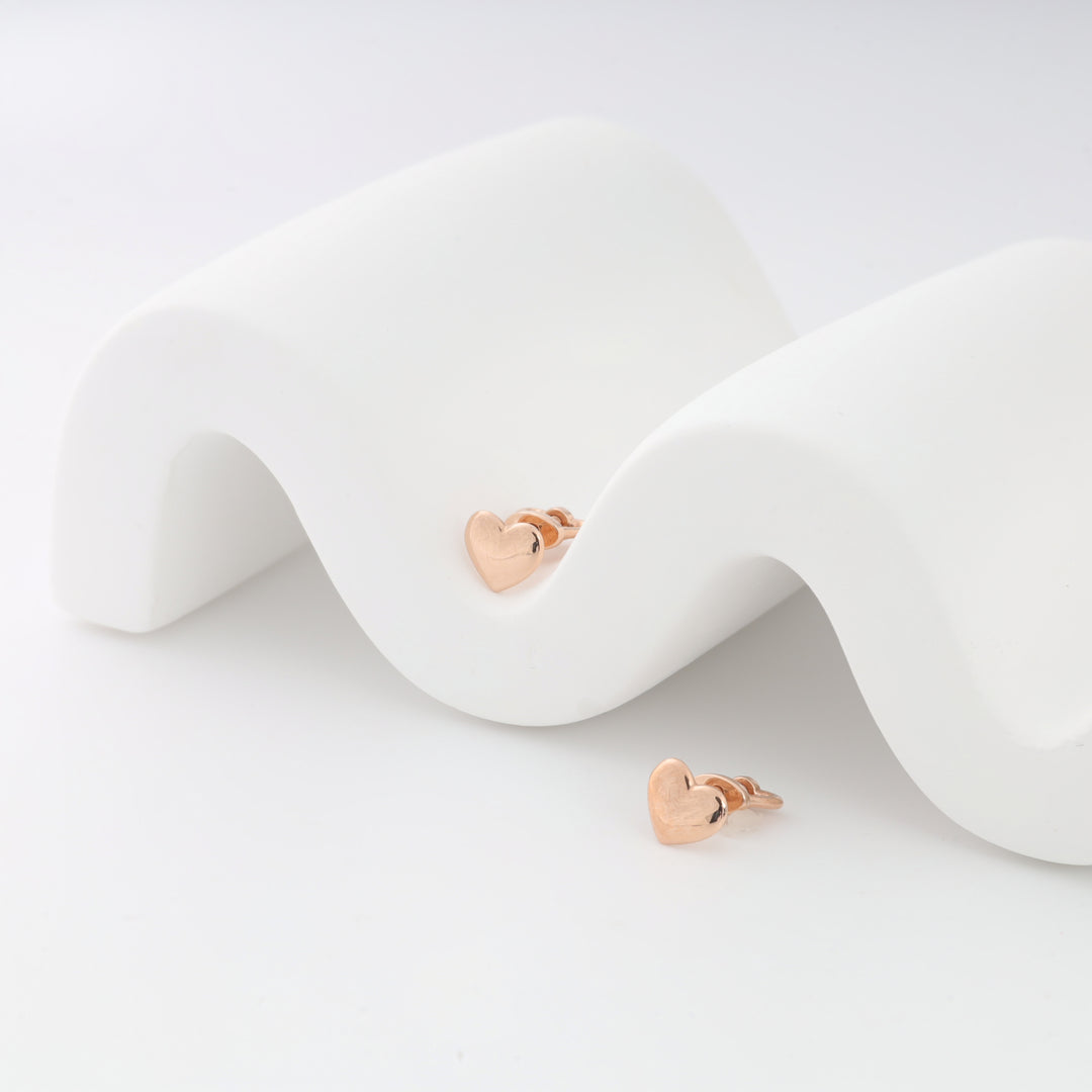 Heart-shaped rose gold stud earrings on a white curved display surface.