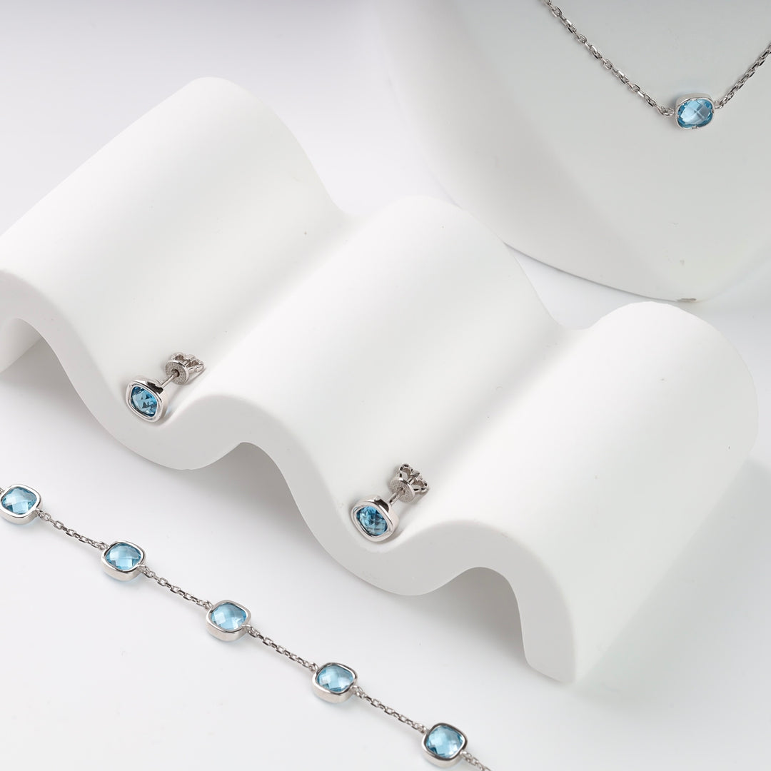 Silver jewelry set featuring blue topaz necklace, bracelet, and earrings displayed on a white surface.