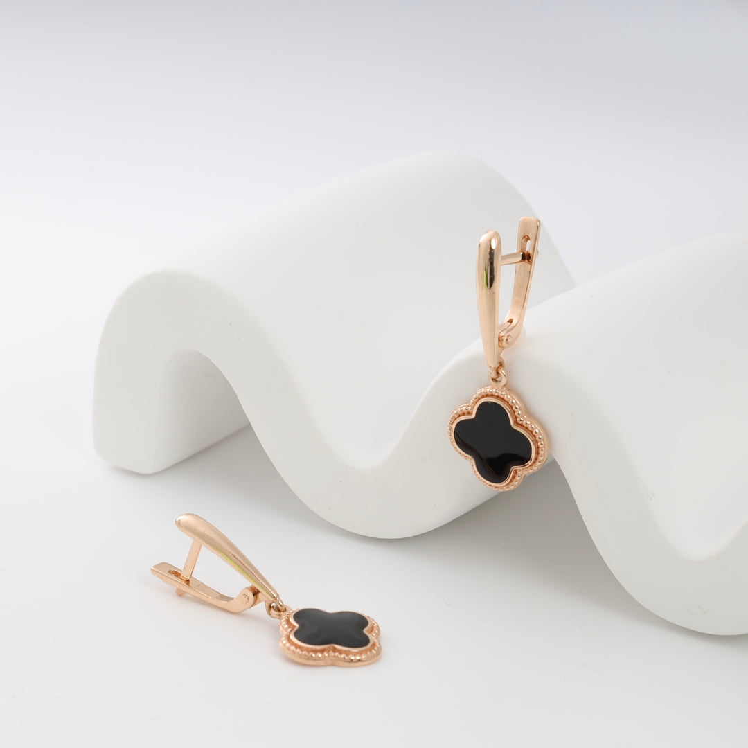 Rose gold earrings with black enamel clover motif on a wavy white background.