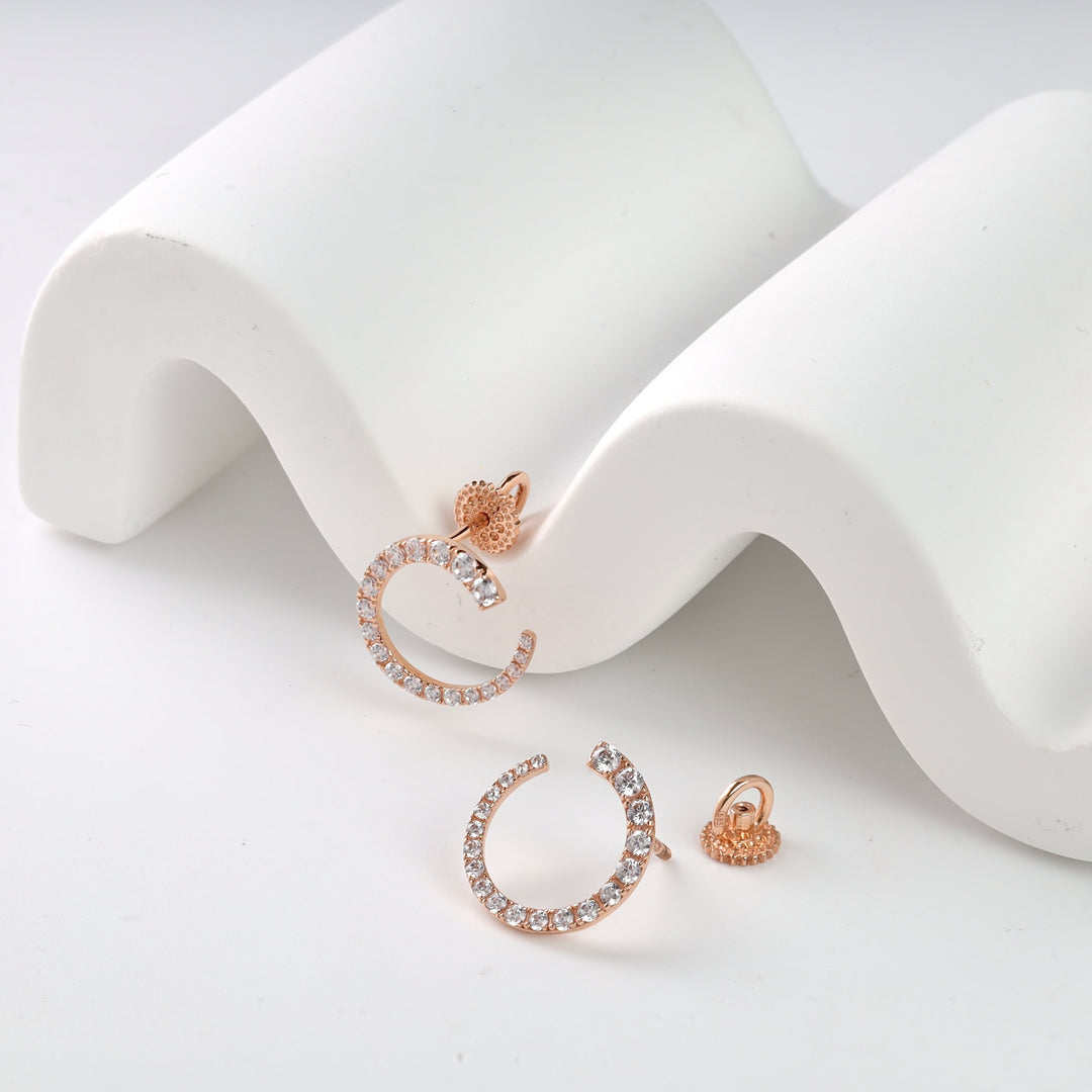 Rose gold crescent-shaped stud earrings with clear crystals on a white wavy surface.