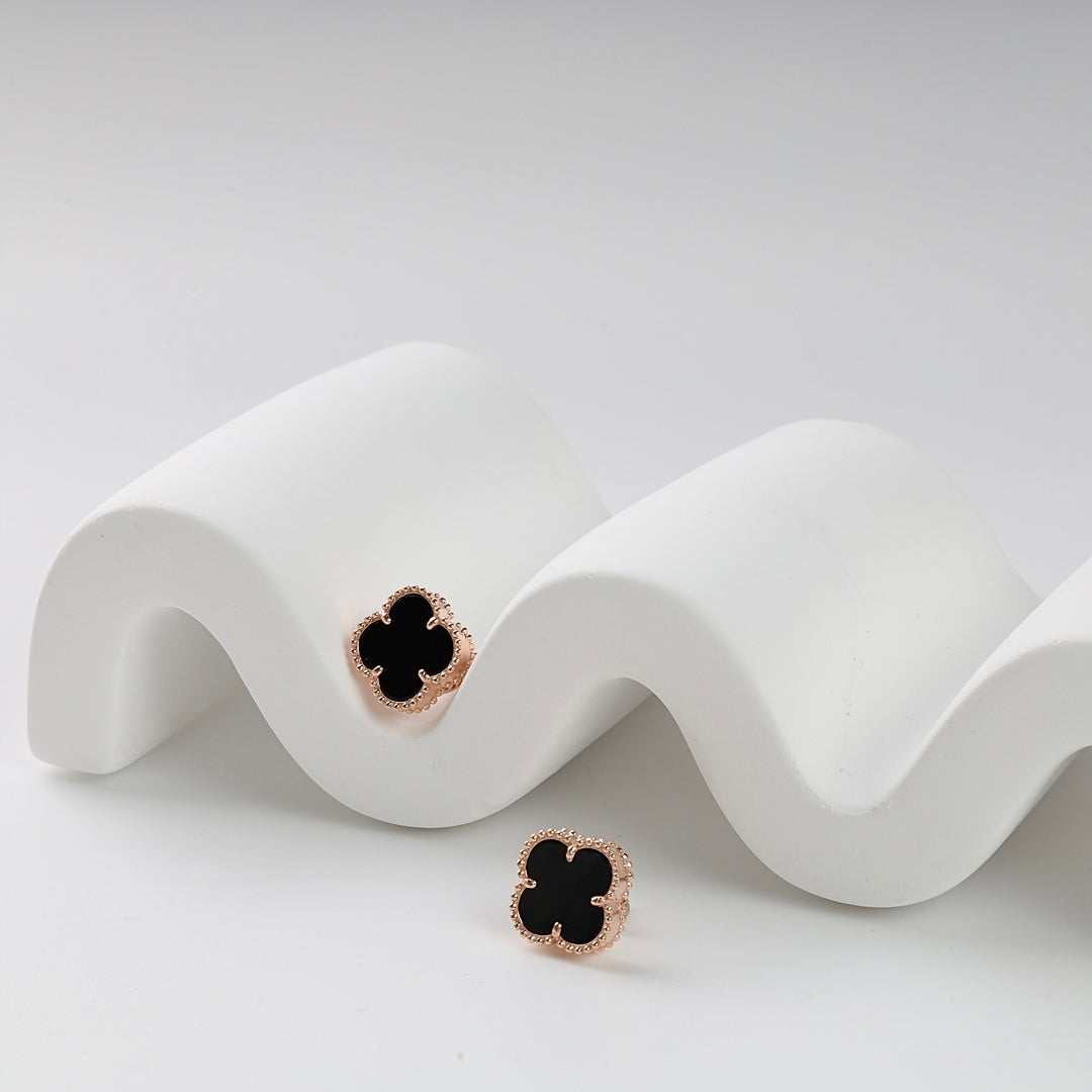 Pair of gold stud earrings with black quatrefoil design displayed on a white wavy stand, modern jewelry, stud earrings.