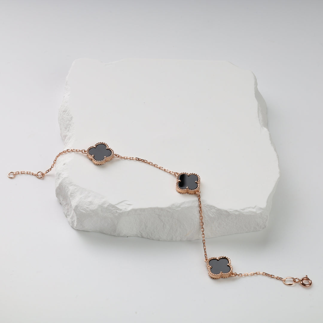 Rose gold chain bracelet with three black clover-shaped charms displayed on a white textured stone.