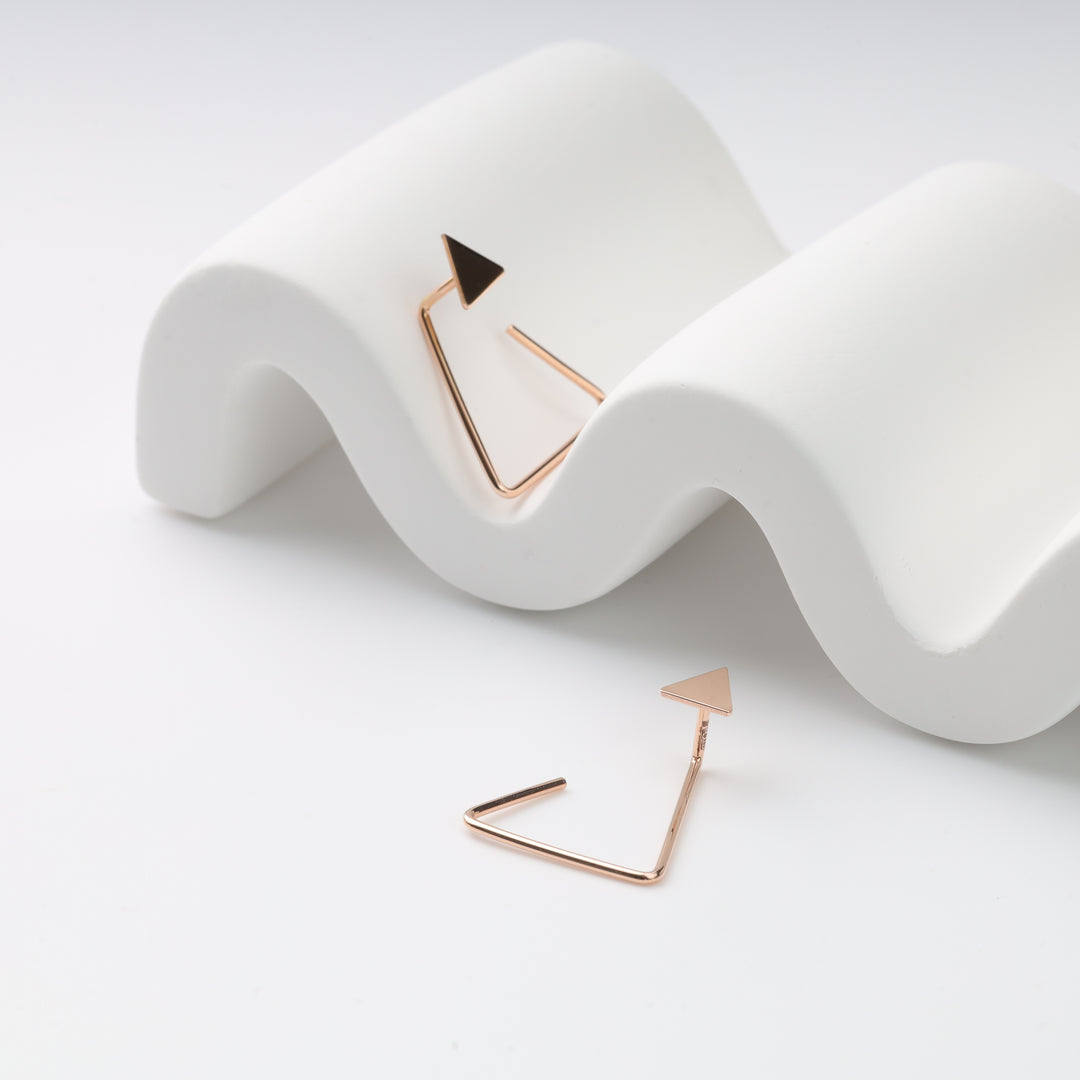 Pair of rose gold triangle earrings, geometric open hoop earrings with triangle studs shown on a white wavy prop.