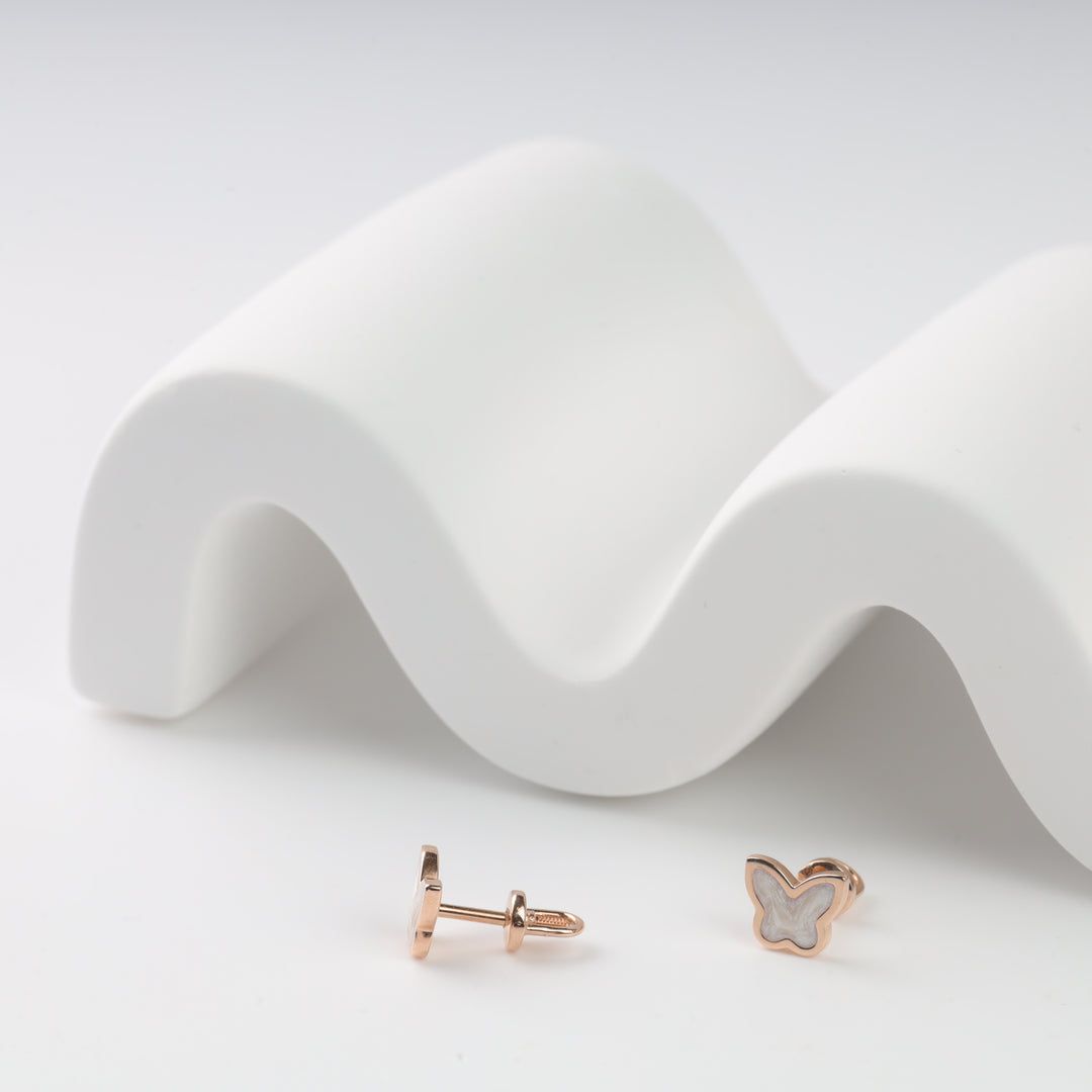 Rose gold and white butterfly stud earrings displayed on a white wavy surface. Perfect for dainty jewelry lovers.