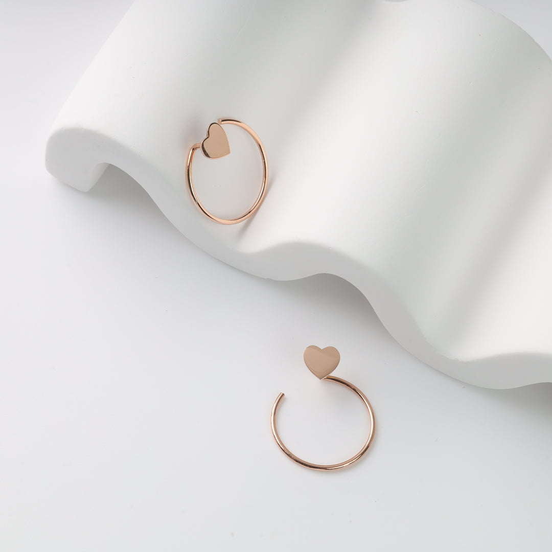 Pair of rose gold open hoop earrings with heart studs displayed on a white wavy stand, rose gold hoop earrings, heart stud hoops.