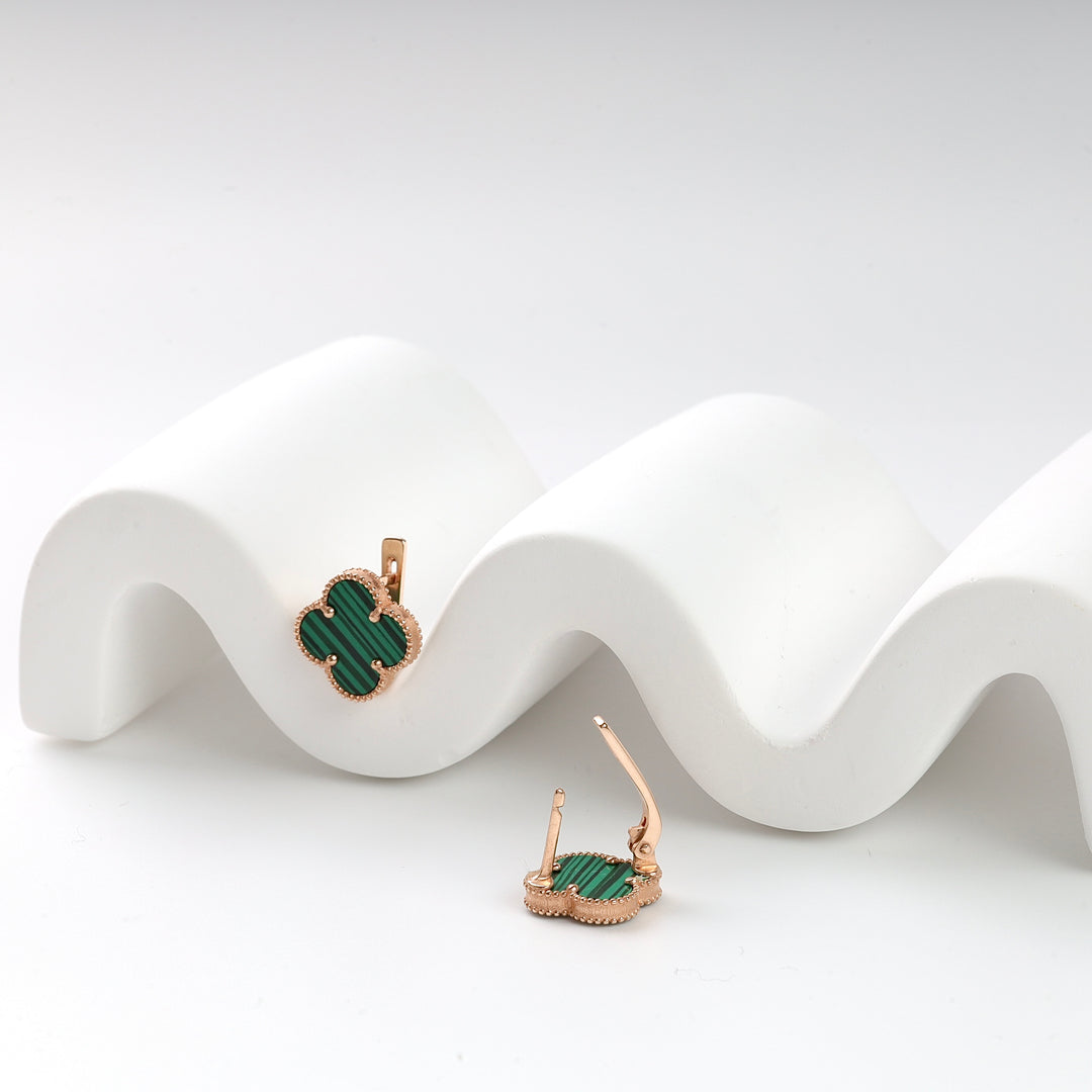 Gold metal clover-shaped earrings with green stone inlay, displayed on a white wavy surface.