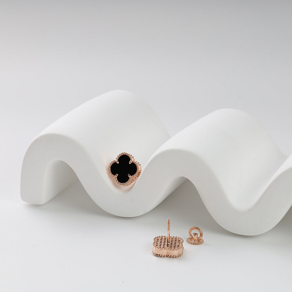 Rose gold quatrefoil pendant with black enamel center and beaded edge displayed on a white wavy surface.