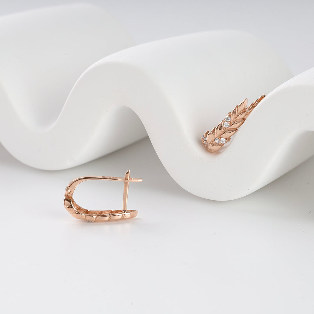 Pair of rose gold hoop earrings with leaf design and small clear stones, displayed on a white wavy surface.