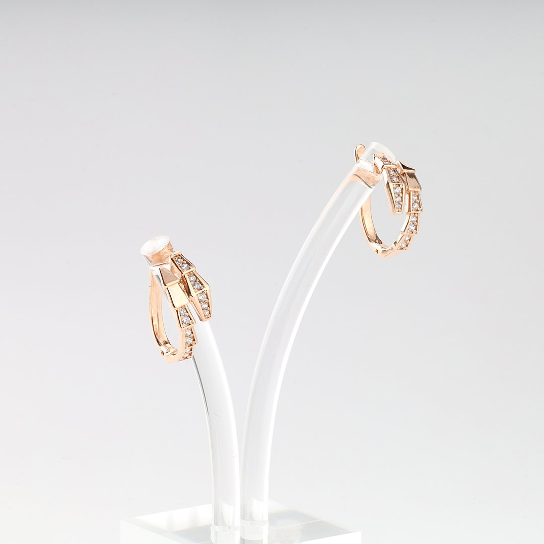 Pair of rose gold hoop earrings with geometric shapes and clear crystals on a white acrylic stand.