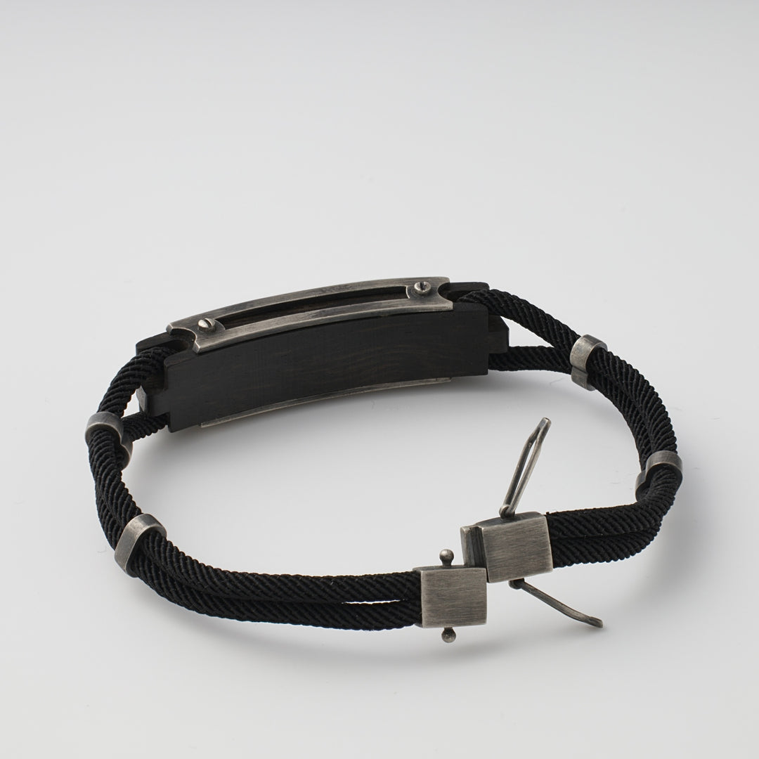 Black cord bracelet with a rectangular oxidized metal plate and silver clasp, displayed on a plain white background.
