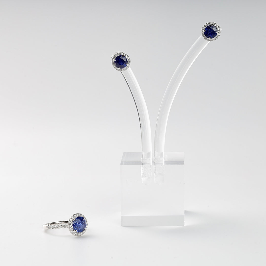 Silver ring and stud earrings set with round blue stones and clear crystal halos, displayed on a white stand.