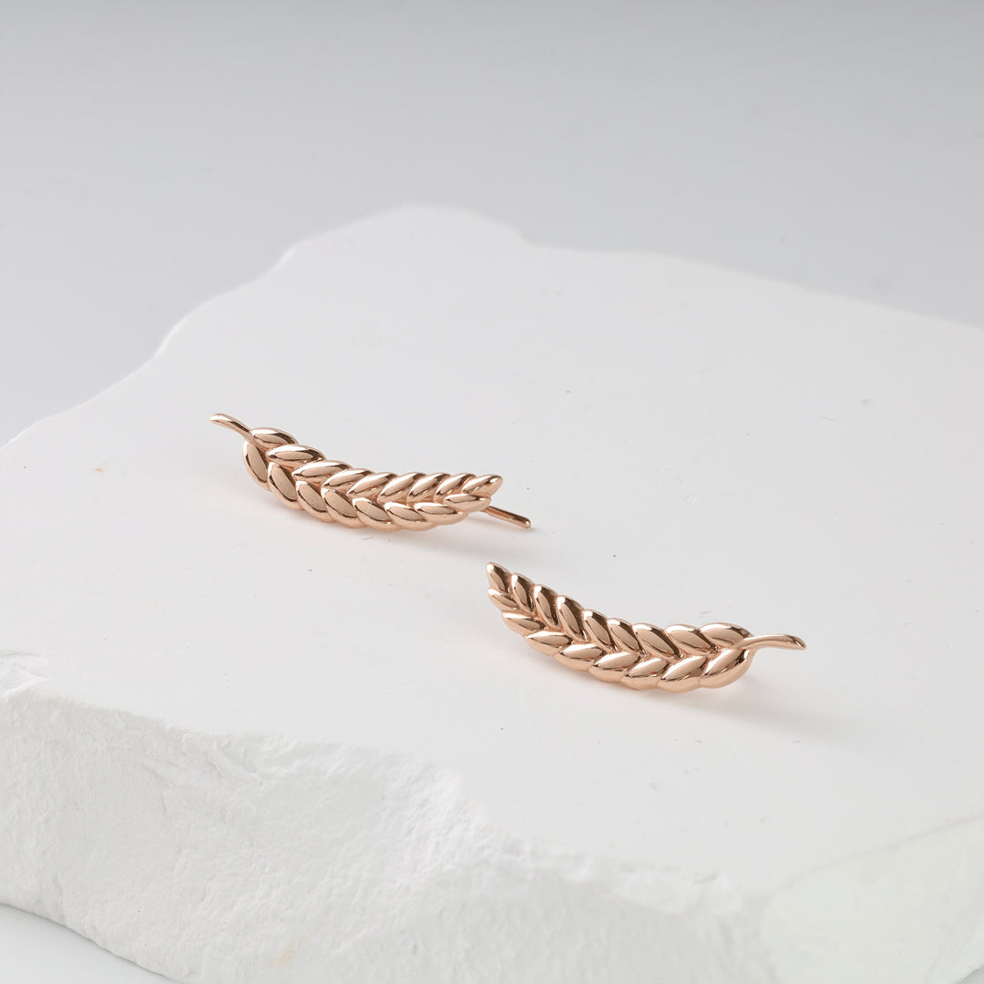 Pair of rose gold leaf motif ear climber earrings displayed on a white textured surface.