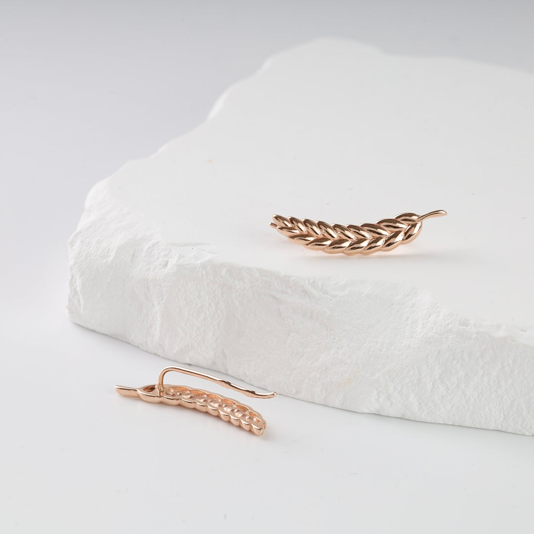 Pair of rose gold ear climber earrings with a leaf design displayed on a white stone surface.