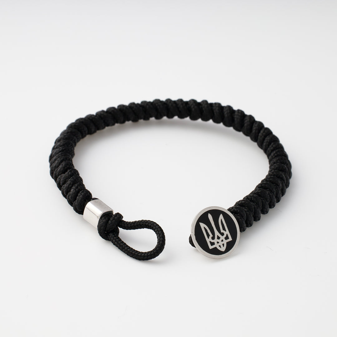 Black braided cord bracelet with a round metal charm engraved with a trident symbol, shown on a white background.