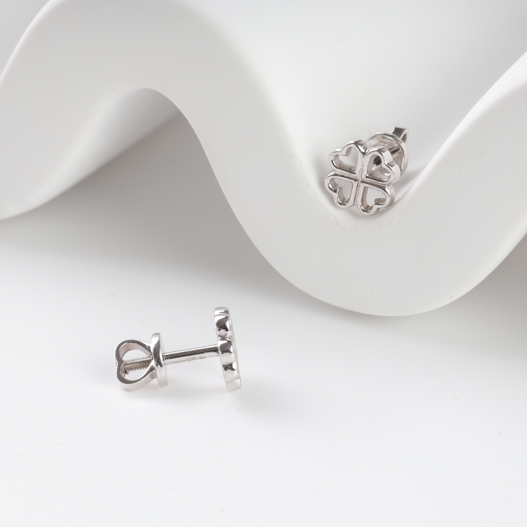 Silver stud earrings in the shape of a four-leaf clover with heart petals, displayed on a white wavy surface.