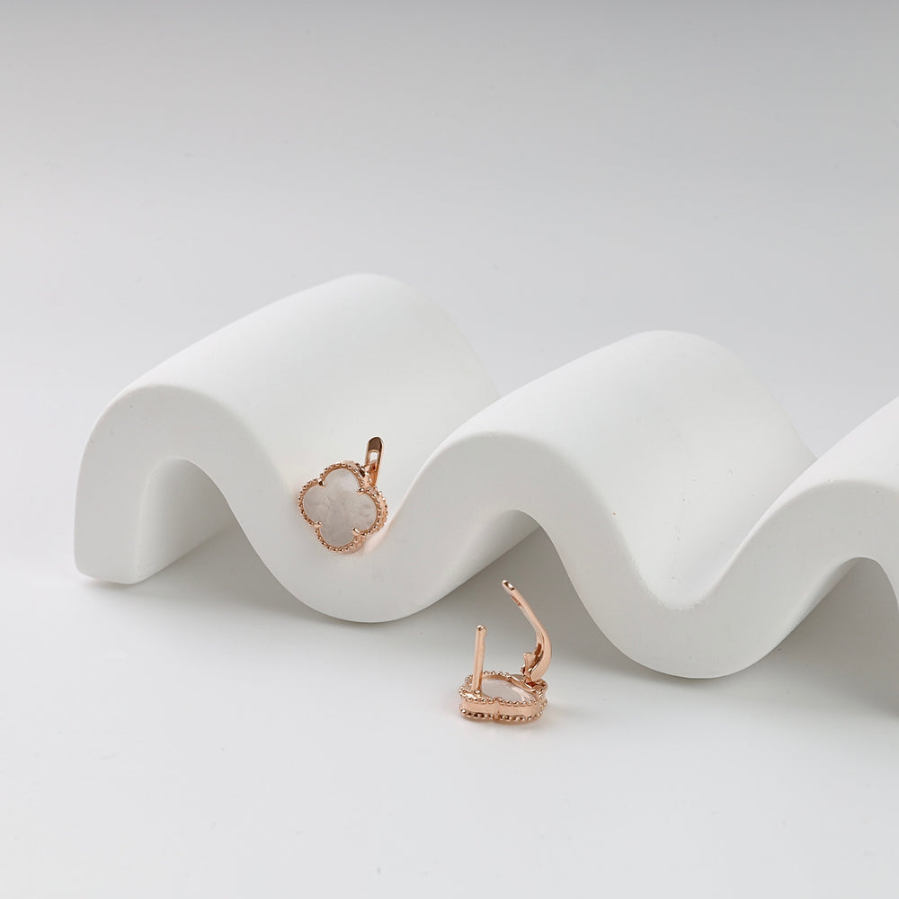 Pair of rose gold clover earrings with white enamel, displayed on a wavy white stand.