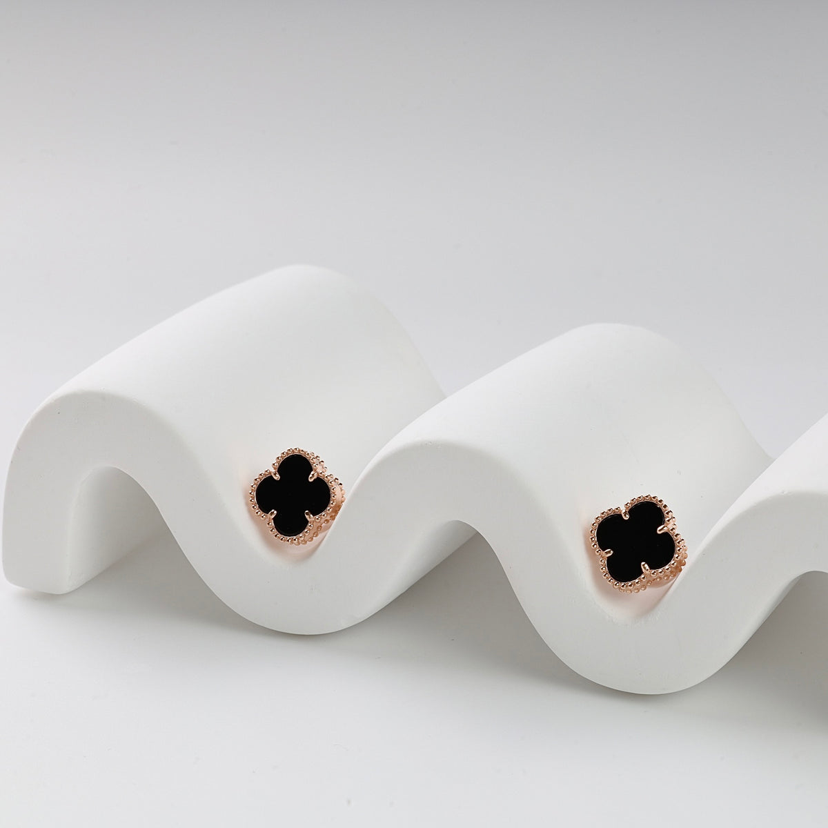 Pair of black enamel four-leaf clover stud earrings with gold trim displayed on a white wavy stand.