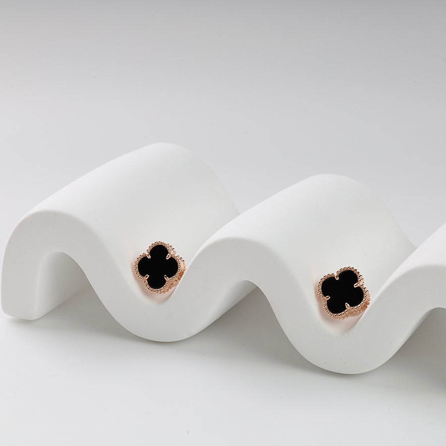 Pair of black enamel four-leaf clover stud earrings with gold trim displayed on a white wavy stand.